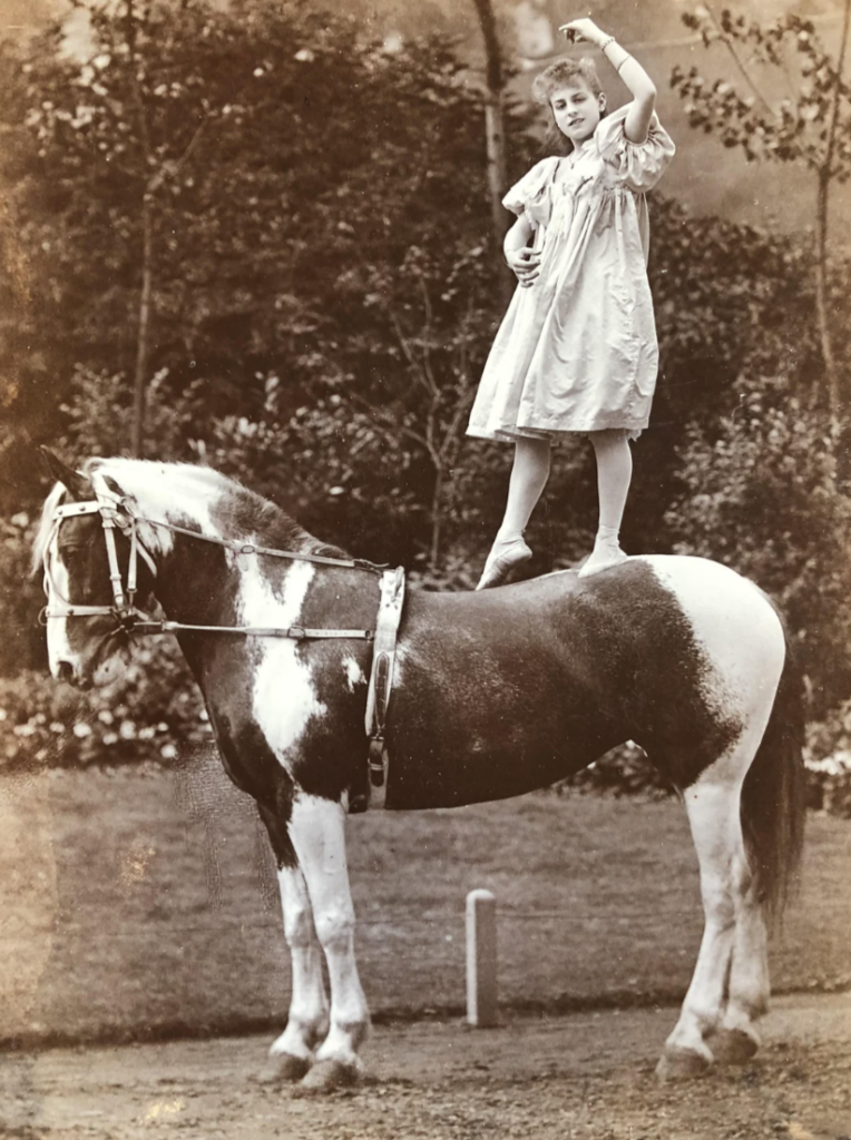 A young girl in a light dress stands on one foot atop a black and white horse in a garden. She raises one arm gracefully above her head. Trees and foliage form the background. The image is in sepia tone.