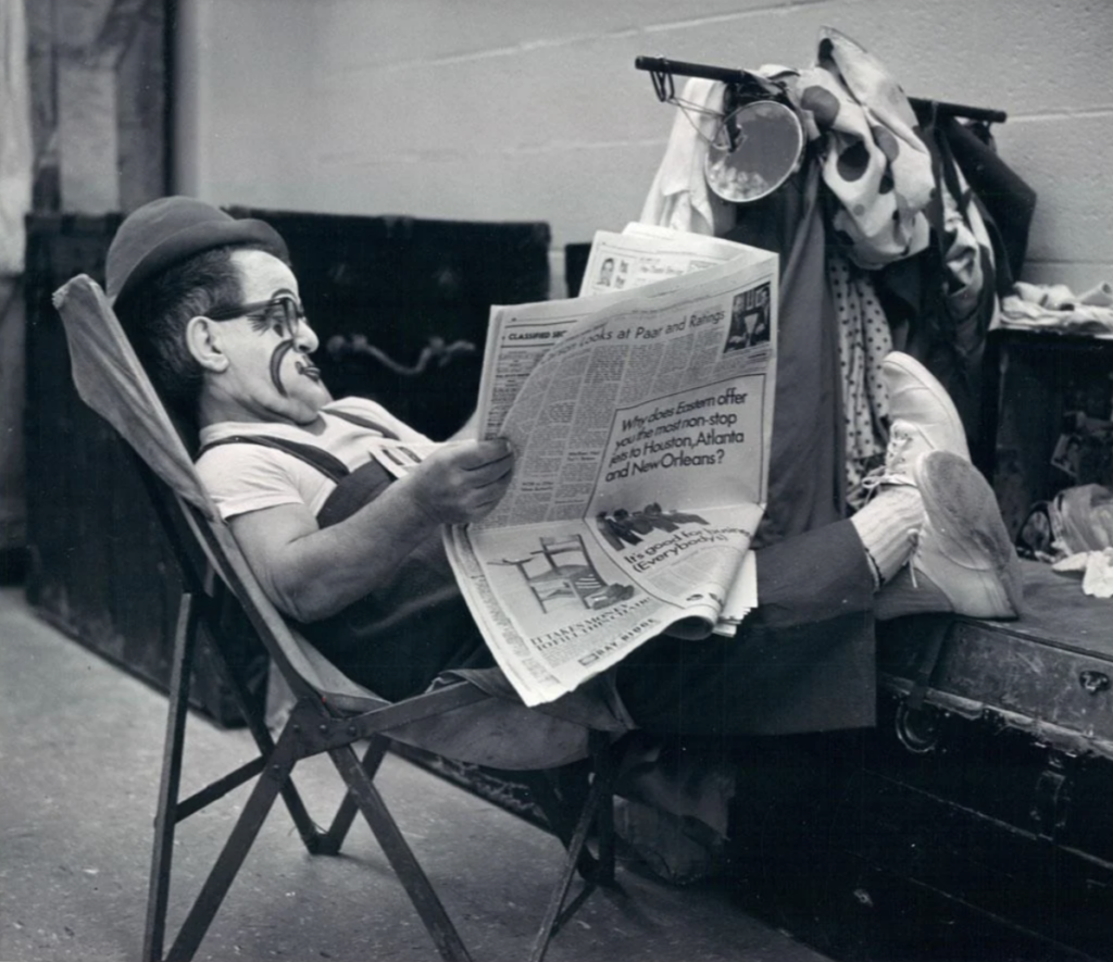 A person dressed as a clown is lounging on a folding chair, reading a newspaper. They are wearing a bowler hat, glasses, clown makeup, and oversized shoes. Clown costumes are hanging nearby on a coat rack. The setting appears to be backstage.