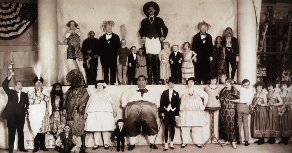 A vintage photo of various circus performers posing on stage. The group includes people of varying heights and body types, dressed in formal attire. Some wear costumes or masks. An American flag is partially visible in the background.