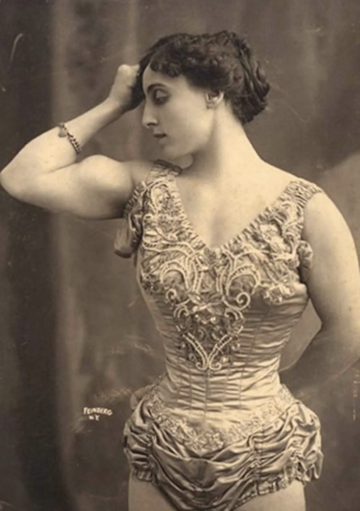 A vintage sepia-toned photograph of a woman in an ornate, embellished costume with intricate designs. She poses with one arm flexed, resting her hand on her head, showcasing her muscular build. Her hair is styled elegantly.