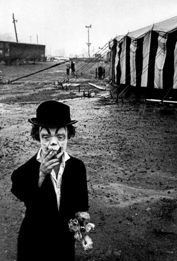 A clown in a suit with face paint smokes a cigarette while holding flowers. He stands on a muddy path leading to a striped circus tent. The background is overcast with people and poles scattered in the distance.