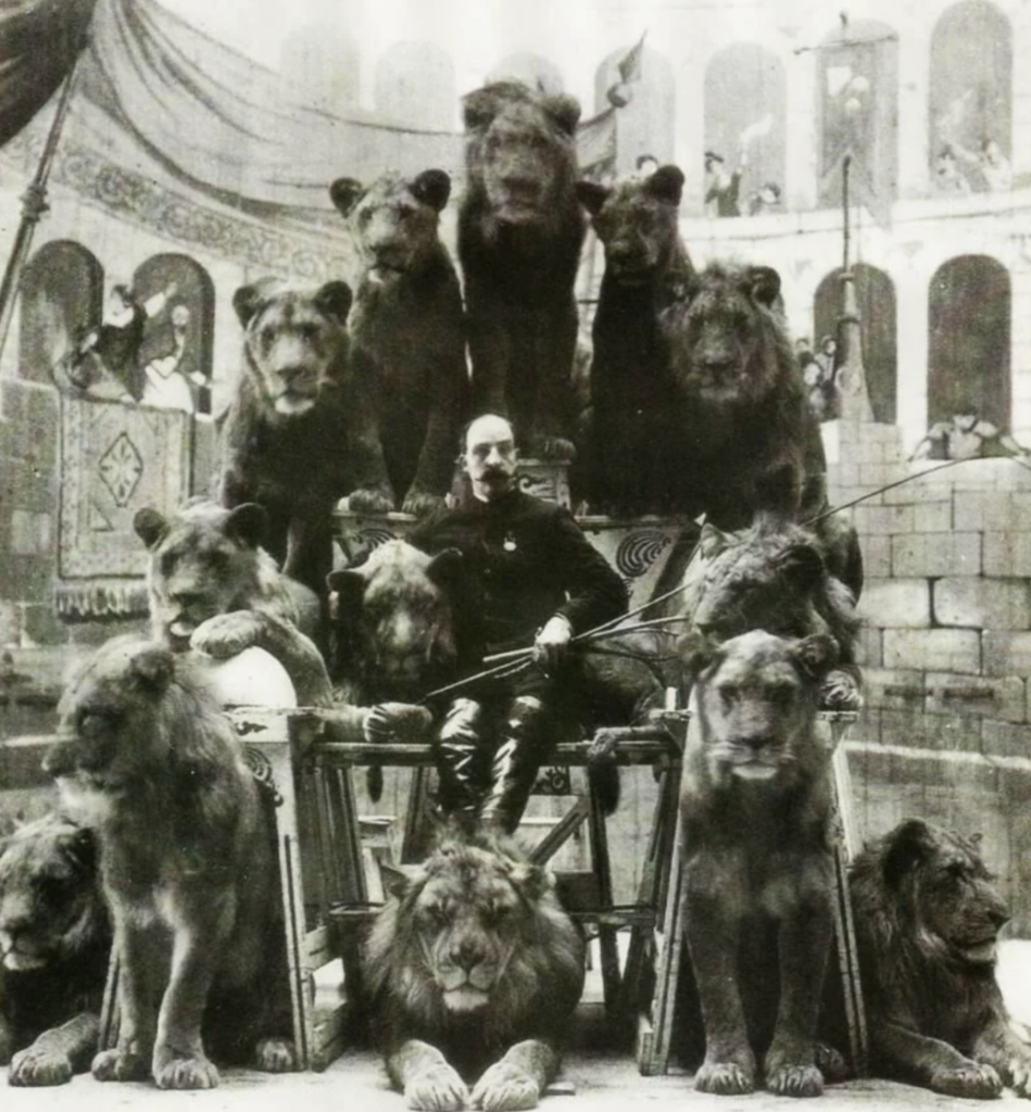 A man sits confidently among a group of lions in an arena with onlookers. The lions are positioned around and above him on platforms. The scene gives an impression of a circus or performance setting with Roman architecture in the background. Black and white.