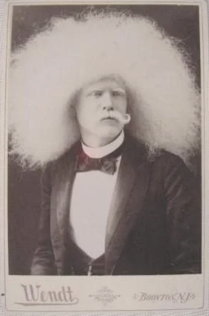 A sepia-toned portrait of a person with a large, fluffy white wig, wearing formal attire with a bow tie. They hold a white pipe in their mouth. The image has "Wendt" and "Boonton, N.J." inscribed at the bottom.