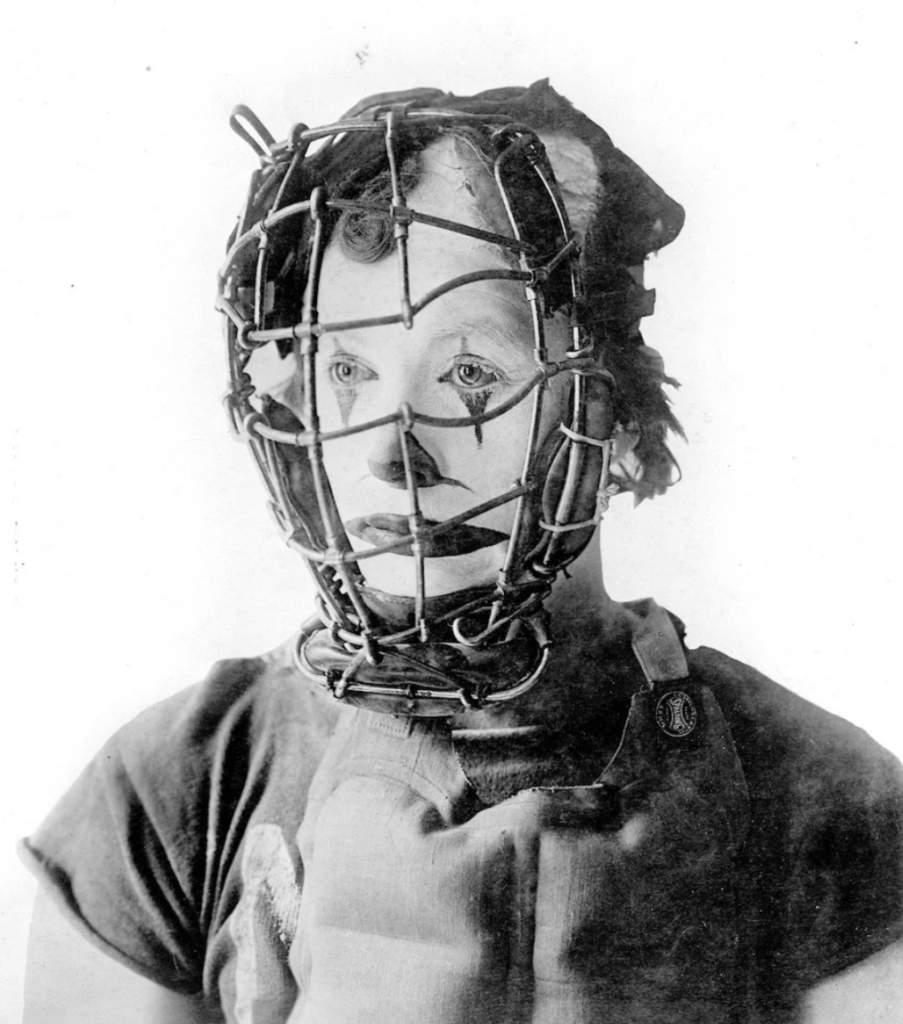 A person wearing a catcher’s mask and a painted clown face, with a short-sleeved top, in a black and white photograph. The expression is neutral.