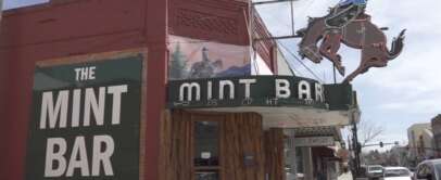 Street view of The Mint Bar, featuring a vintage sign with a cowboy on a bucking horse. A green sign on the brick facade reads, "The Mint Bar Since 1907 'Where Good Friends Meet'." Nearby stores and a sidewalk are visible.