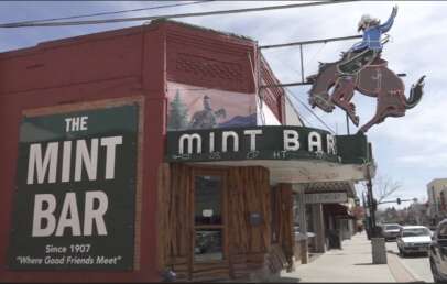 Street view of The Mint Bar, featuring a vintage sign with a cowboy on a bucking horse. A green sign on the brick facade reads, "The Mint Bar Since 1907 'Where Good Friends Meet'." Nearby stores and a sidewalk are visible.