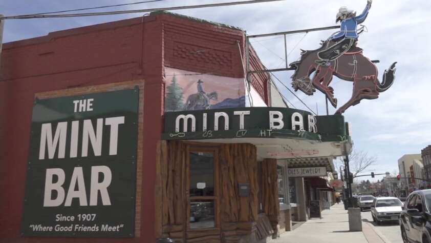 Street view of The Mint Bar, featuring a vintage sign with a cowboy on a bucking horse. A green sign on the brick facade reads, "The Mint Bar Since 1907 'Where Good Friends Meet'." Nearby stores and a sidewalk are visible.