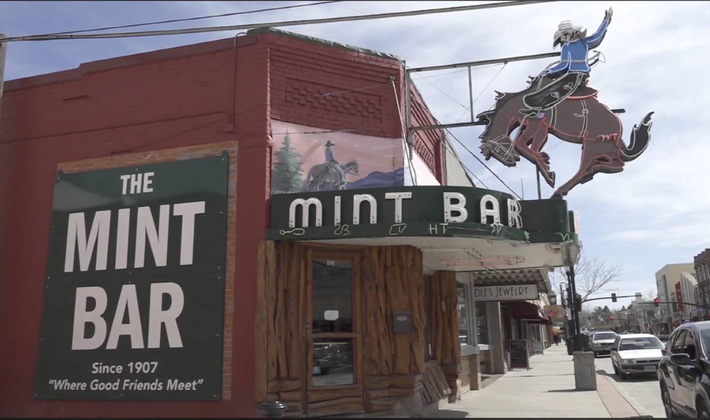 Street view of The Mint Bar, featuring a vintage sign with a cowboy on a bucking horse. A green sign on the brick facade reads, "The Mint Bar Since 1907 'Where Good Friends Meet'." Nearby stores and a sidewalk are visible.