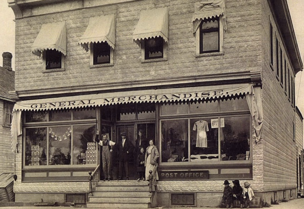 A vintage general merchandise store with an overhead sign and awnings. Five people stand on the steps, and a dog sits nearby. Clothing is displayed in the windows. The building also functions as a post office, visible in the signage.