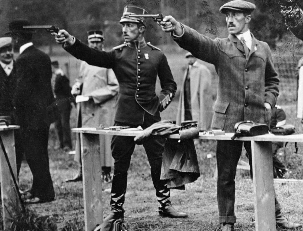 Two men participating in an early 20th-century pistol shooting competition. One is in a military uniform, and the other wears a suit and flat cap. Both are aiming pistols at targets. Observers stand in the background.