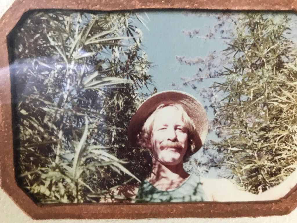 A person in a straw hat and tank top smiles while standing among tall plants under a clear blue sky. The photo has a vintage look with a brown-bordered edge.