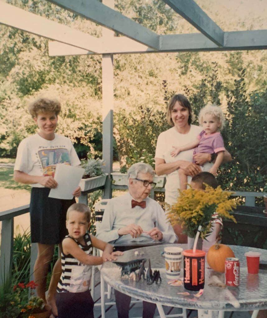 A group of people on a patio: an older man in a bow tie sits at a table with a dinosaur set; kids surround him; two women, one holding a child, are nearby. The table has flowers and soda cans. The background is lush with greenery.