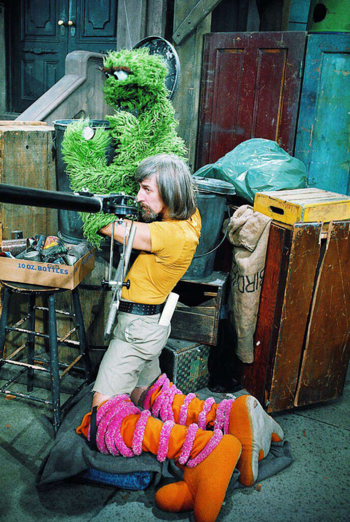 A puppeteer sitting on a crate, operating a large green furry puppet with a pipe behind it. Next to him is an orange furry puppet laying on the floor. Various wooden boxes and bags are in the background.