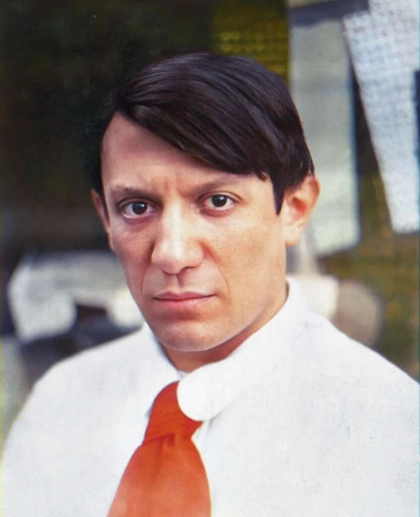 A person with short, dark hair wearing a white shirt and a bright red tie. The background is blurred with soft colors.