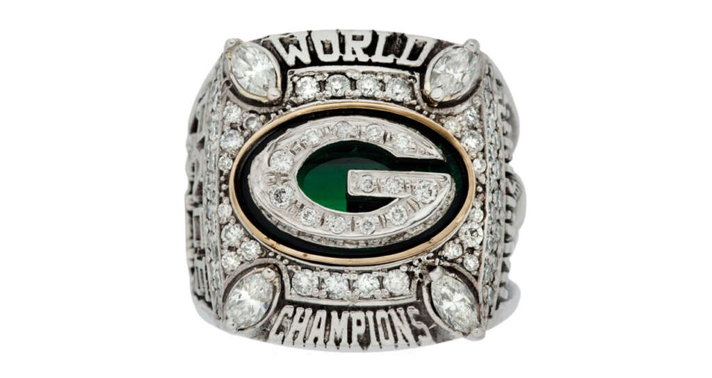 A championship ring with a prominent "G" logo at the center, surrounded by numerous small diamonds. The words "World Champions" are engraved on the top and bottom, with additional decorative elements around the band.