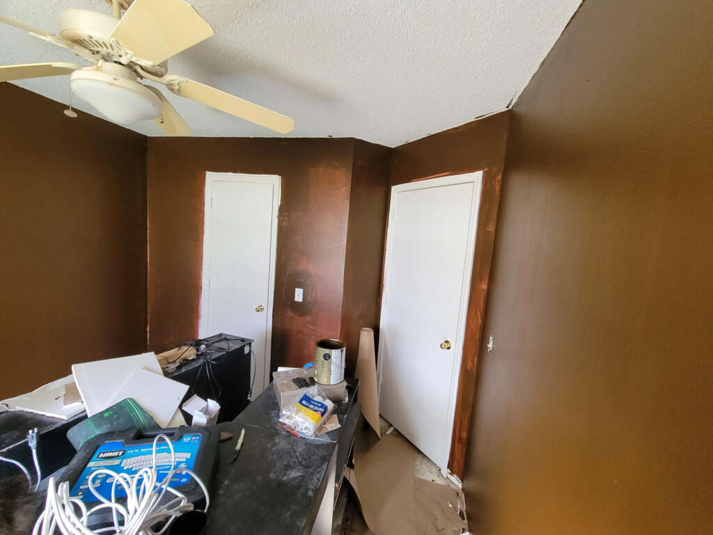 A room with brown walls is undergoing renovation. Two doors are visible, partially painted white. The ceiling fan is off. There's clutter, including a can of paint, cables, and various items scattered on a black surface.