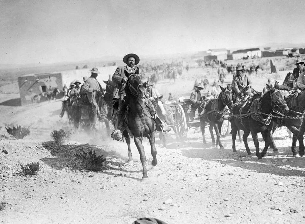 Historical black and white photo of a man on horseback leading a group of soldiers. The scene is set in a desert area with several buildings in the background and soldiers on horseback and carriages following behind, raising dust.