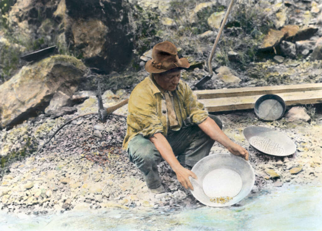 A person in a brown hat and yellow shirt crouches by a river, panning for gold. The scene includes a metal pan with water and sediment, surrounded by rocks and greenery, illustrating a historical gold mining activity.