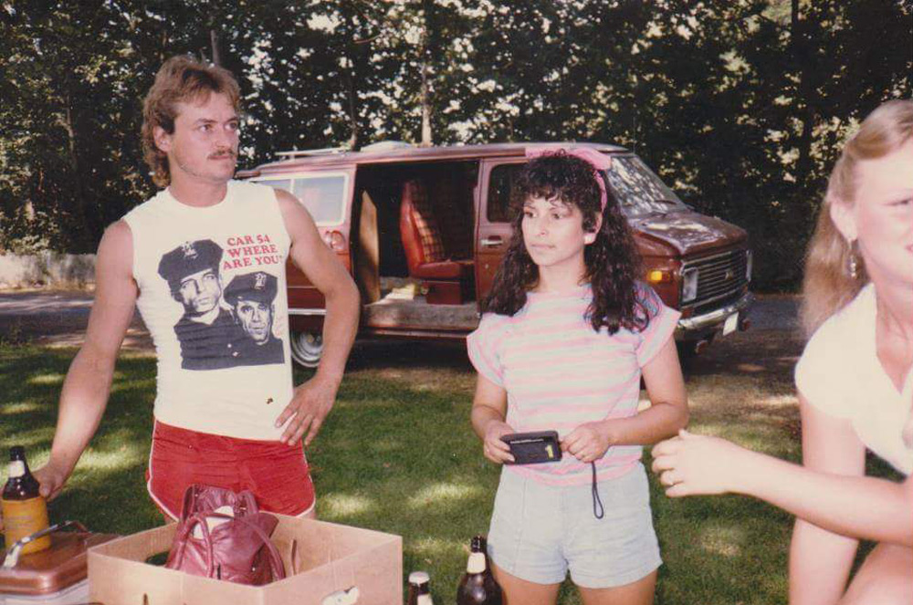 A man in a sleeveless shirt with a graphic and red shorts stands next to a woman holding a small object. She wears a pink striped top and denim shorts. A red van is parked in the background. There's a table with bottles and bags in the foreground.