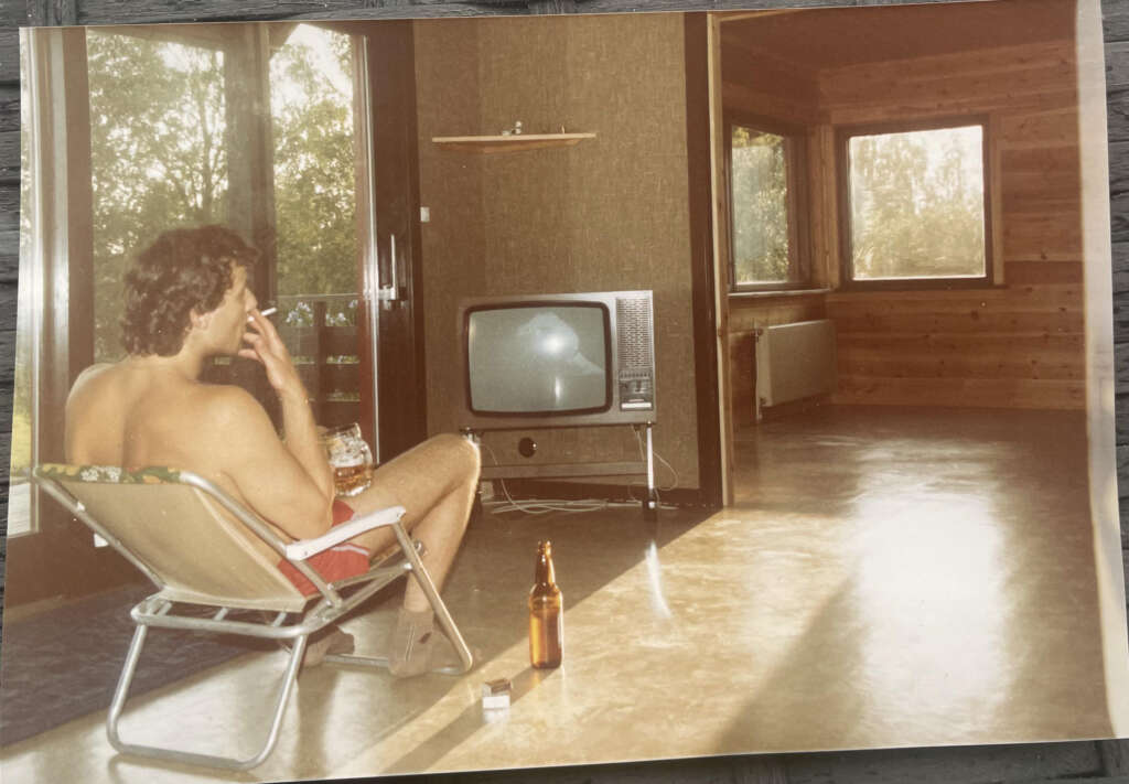 A person sitting on a lawn chair indoors, holding a drink and looking at a vintage TV. An empty beer bottle and a pack of cigarettes are on the floor. The room has wooden walls, a large window, and a sliding door letting in natural light.