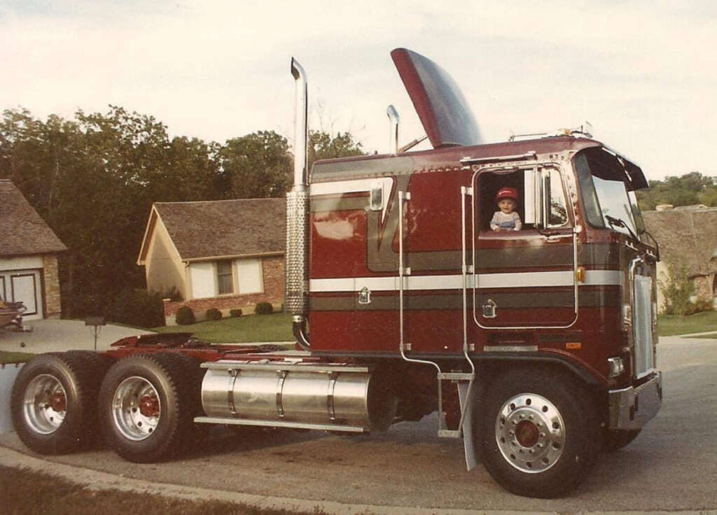 A child wearing a hat is sitting inside a red and gray semi-truck with a raised cab roof, parked on a suburban street. The background features houses and trees under a cloudy sky.