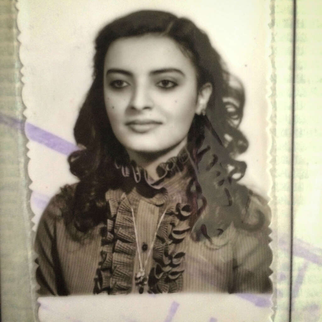 Black and white portrait of a woman with long, dark, wavy hair wearing a ruffled blouse. She looks away from the camera with a slight smile. The image has a vintage, textured appearance.