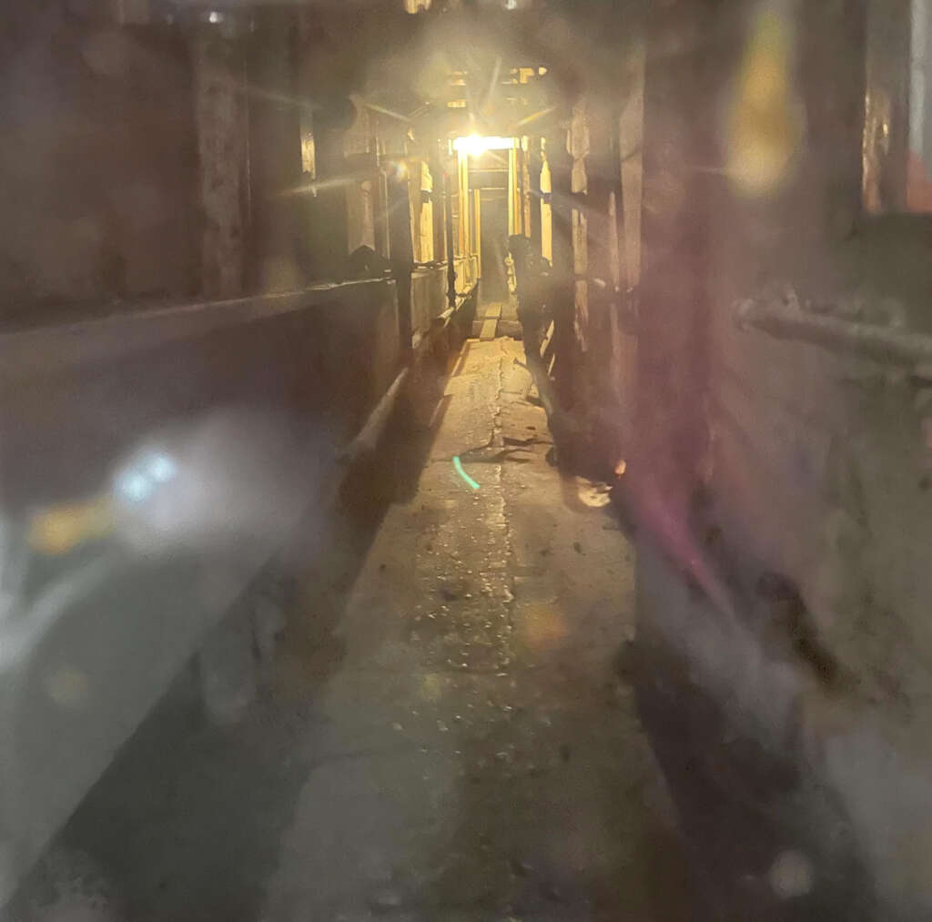 Dimly lit narrow alleyway with a glowing light at the end. The path is surrounded by walls and faint structures, creating a shadowy and mysterious atmosphere. Dust particles are visible in the air, adding to the eerie ambiance.