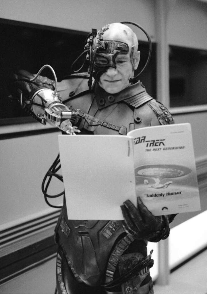 A person in an elaborate sci-fi costume, featuring mechanical components on their head, is holding a script labeled "Star Trek: The Next Generation - Suddenly Human." They appear to be on a set, reading the script attentively.