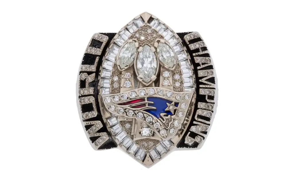 A championship ring featuring the New England Patriots logo, surrounded by numerous sparkling diamonds and the words "World Champions." The intricate design includes marquise and baguette-cut stones.