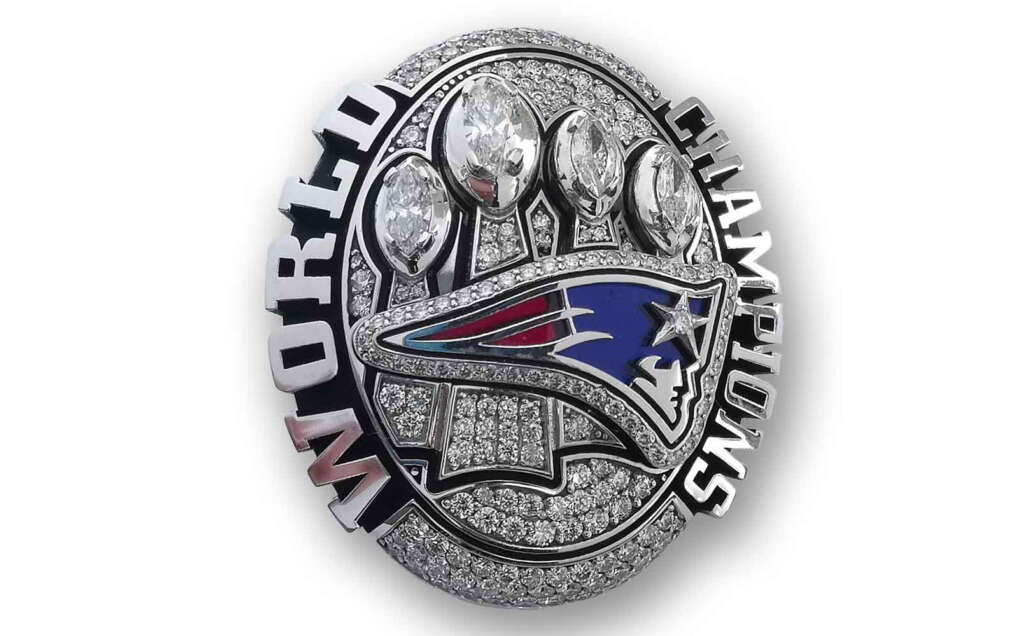 A championship ring with "World Champions" inscribed, featuring a sparkling design with five football-shaped diamonds and a logo resembling a stylized patriot.