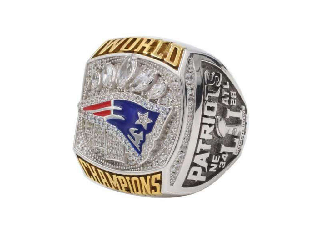 A championship ring with "World Champions" engraved. It has the logo of a sports team adorned with diamonds. The side details "PATRIOTS" and the scores "NE 34, ATL 28".