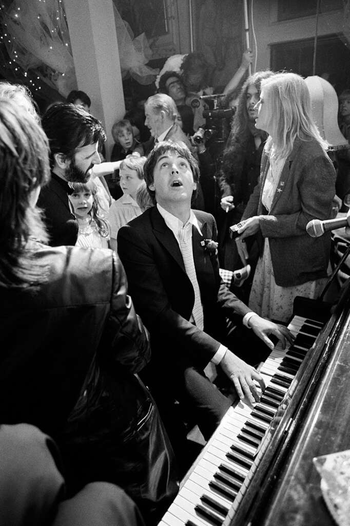 Black and white photo of a man playing a piano at a lively gathering. He wears a suit and looks upwards while others, including a person with a camera and people in casual clothing, surround him, creating a festive atmosphere.