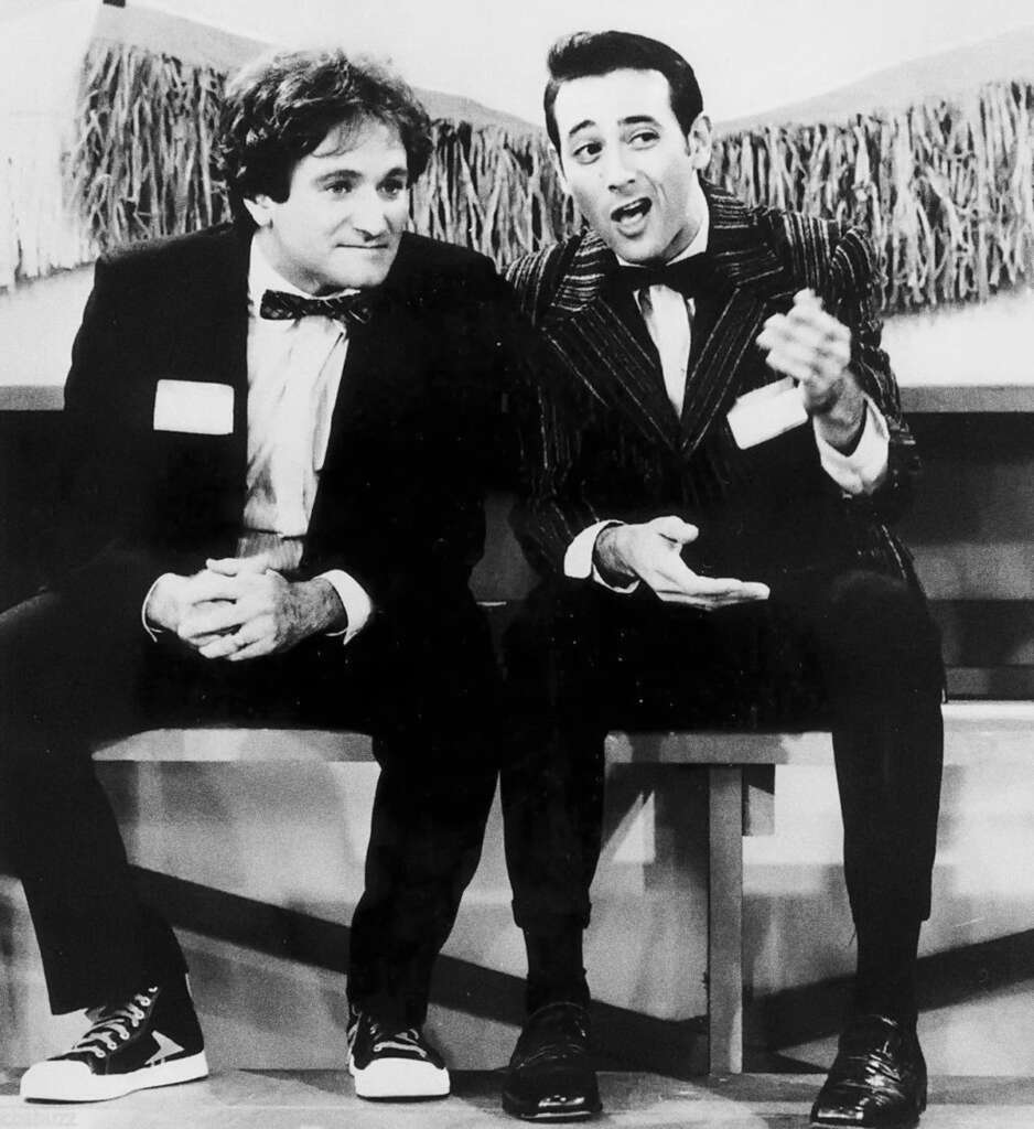 Two men sit on a bench wearing suits; the one on the left is in sneakers, and the one on the right gestures animatedly. Behind them is fringed decor, suggesting a playful or casual setting. They both wear name tags.