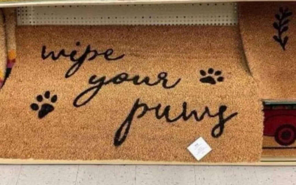 A doormat with the phrase "wipe your paws" written in black cursive. There are paw prints on either side of the text, and the mat is displayed on a store shelf.