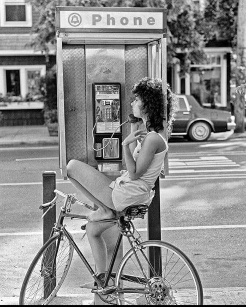 A woman with curly hair sits on a bicycle, holding a phone receiver at an outdoor phone booth. She is wearing a tank top and shorts, and the scene is set on a street with parked cars and houses in the background. The photo is in black and white.