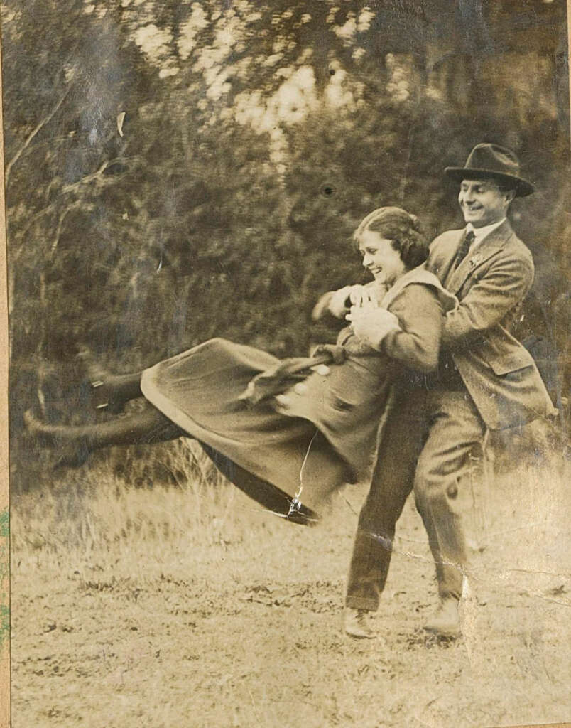 A sepia-toned vintage photo shows a man joyfully swinging a smiling woman in a long coat. They are outdoors in a park-like setting, surrounded by blurred trees and grass, both appearing happy and playful.