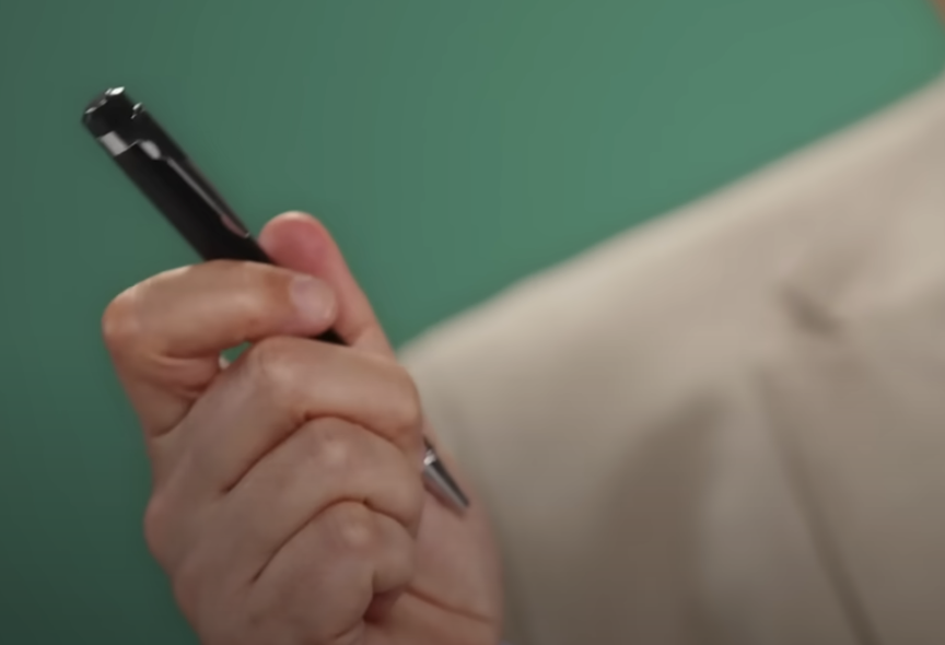 A person holding a black pen with a silver tip and clip, against a blurred green and beige background. Their thumb and fingers are visible, gently gripping the pen.