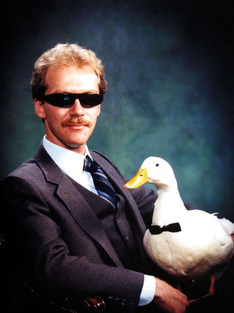 A man in a suit with sunglasses and a mustache sits against a dark green background, holding a duck wearing a bow tie.