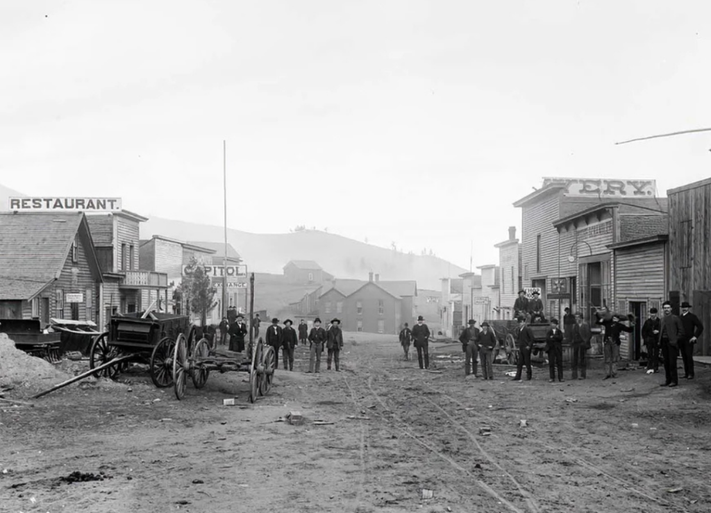 A vintage black and white photo of a dusty street in a frontier town, with wooden buildings labeled "RESTAURANT" and "CAPITOL." People and horse-drawn carts are gathered along the road. Hills are visible in the background.