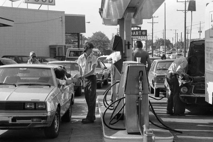 Black and white photo of a busy 1970s gas station. Attendants are refueling cars and checking engines. Several vehicles are lined up, and a "Mart" sign is visible in the background.