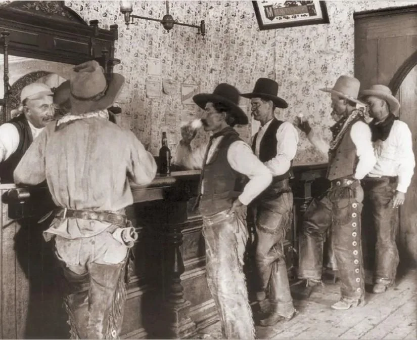 A sepia-toned image of six men in old-fashioned Western attire standing at a wooden bar in a saloon. They are wearing hats, vests, and chaps. The bartender, behind the counter, serves drinks. The room has patterned wallpaper and a framed picture.