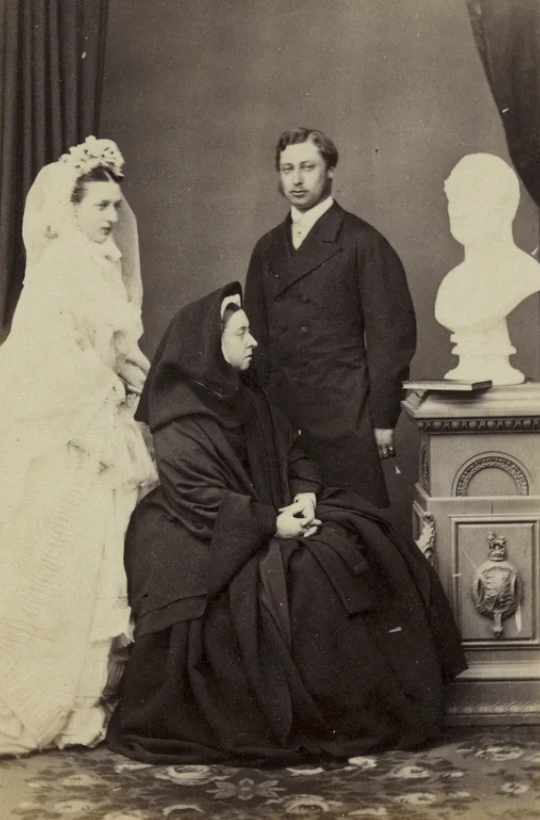 A historical black and white photo featuring three people in formal attire. Two are standing: a woman in a wedding gown and a man in a suit. A seated woman in dark, flowing clothing is beside a bust on a pedestal. They are in an elegantly draped room.