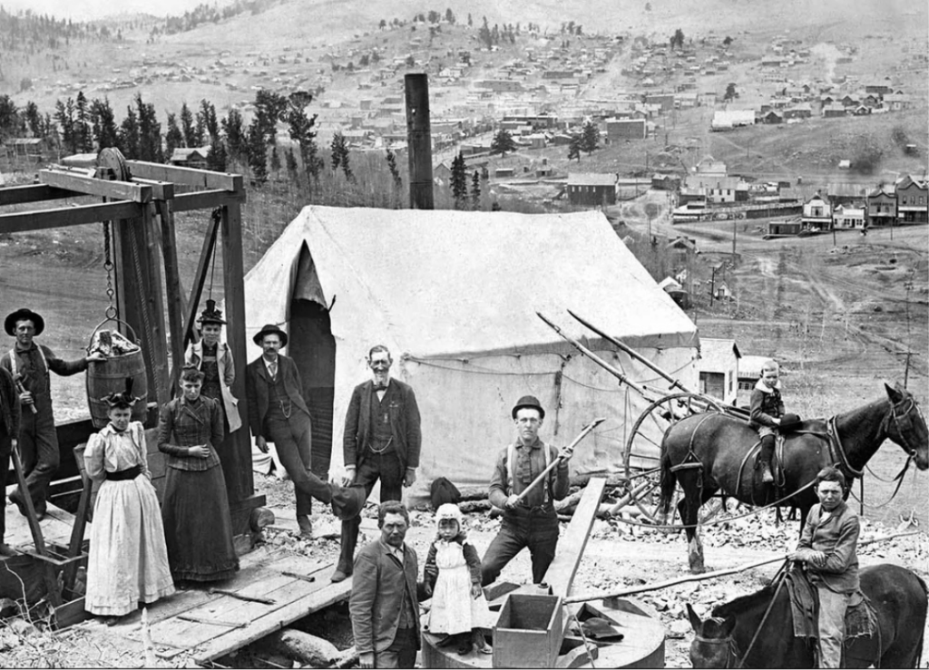 Historical black and white photo shows a group of miners and their families posing in front of a mining camp. A tent and wooden structures are visible, with a mule-drawn cart. A town and hills are seen in the background.