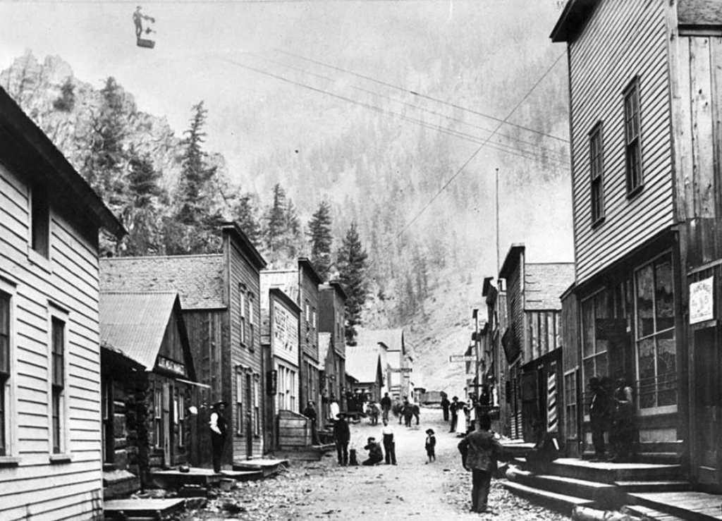 A vintage photo of a narrow street in a rustic town surrounded by mountains. Wooden buildings line both sides of the street. People, including children, are scattered along the street, and a cable car is visible overhead.