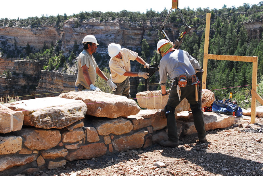 Three construction workers in hard hats are positioning large stones on a wall at a mountainous site. One worker operates a crane, while the others guide the stones into place. A forested canyon is visible in the background.