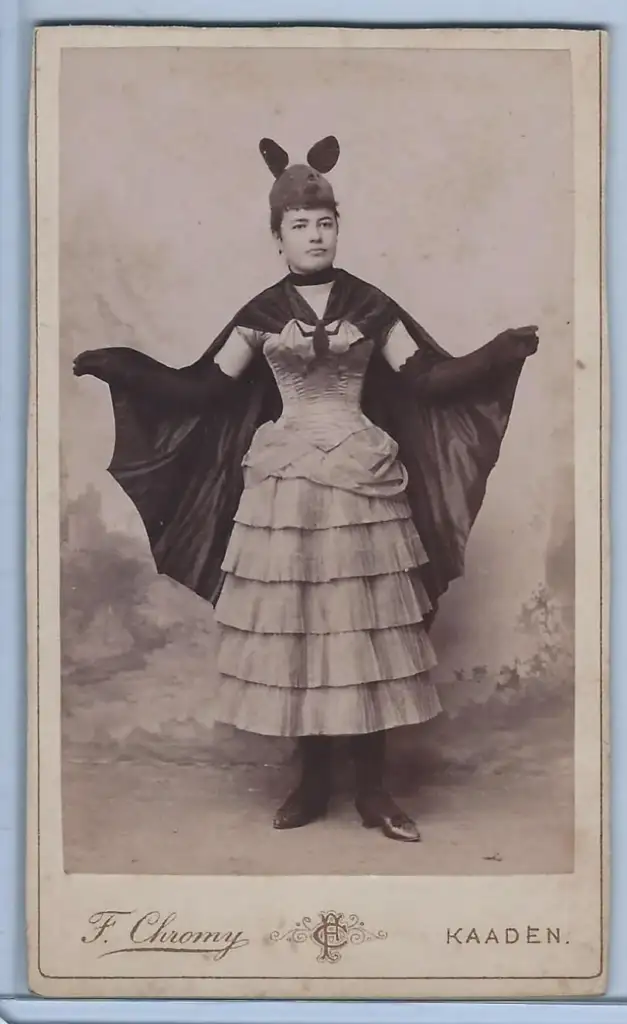 Sepia-toned vintage photograph of a woman wearing a tiered dress and a cape with a bat-like collar. She also has a headpiece with two upward-pointing ears. The background is a faint, artistic rendering.