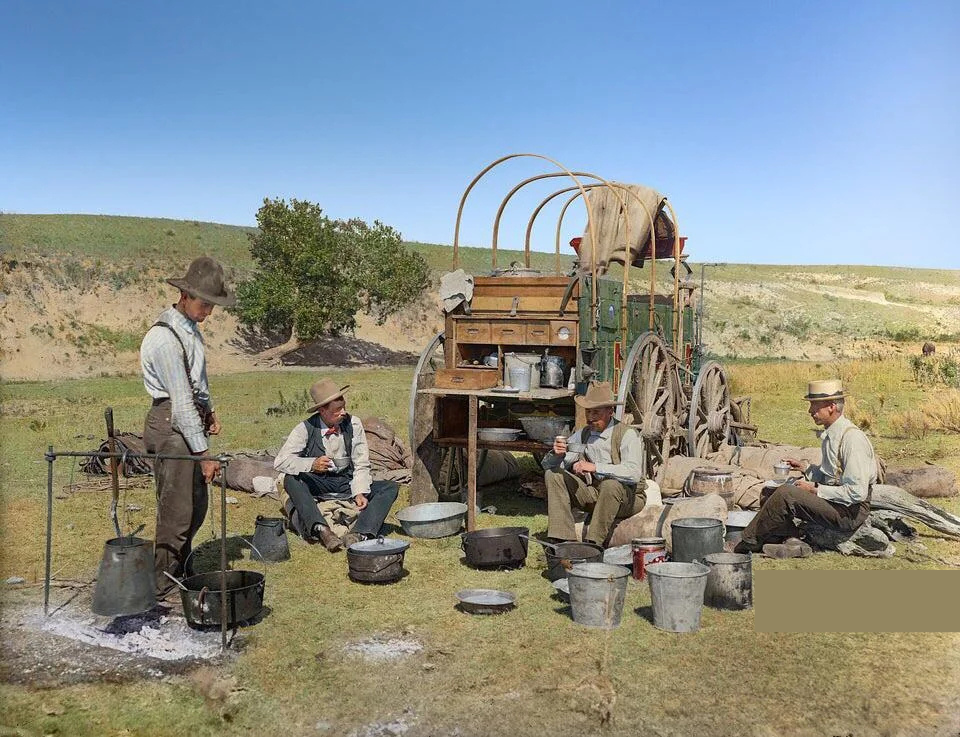 Four men in historical attire sit and stand near a chuckwagon, surrounded by cooking pots. The wagon has a canvas cover and is stationed on grassy terrain with a small hill in the background. The scene suggests an outdoor meal preparation.