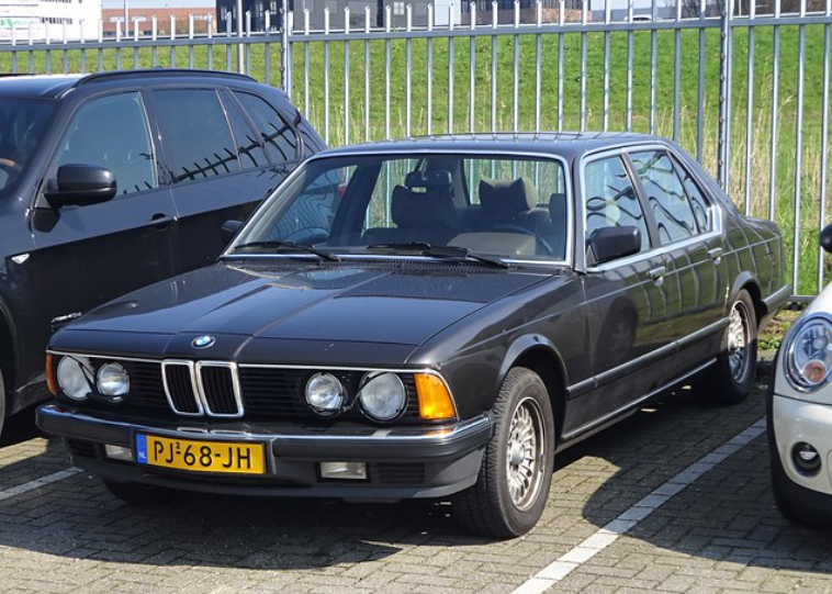 A classic black BMW sedan parked in a parking lot, positioned between two other vehicles. The car features a yellow license plate with the code "PJ-68-JH." A metal fence and a green grassy area are visible in the background.