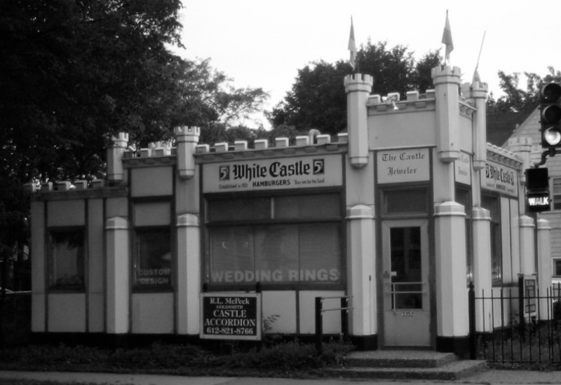 A vintage, castle-themed building with turrets and a sign reading "White Castle Hamburgers" and "The Castle Jeweler." Other signs advertise wedding rings, custom design, and accordions. Trees surround the structure.