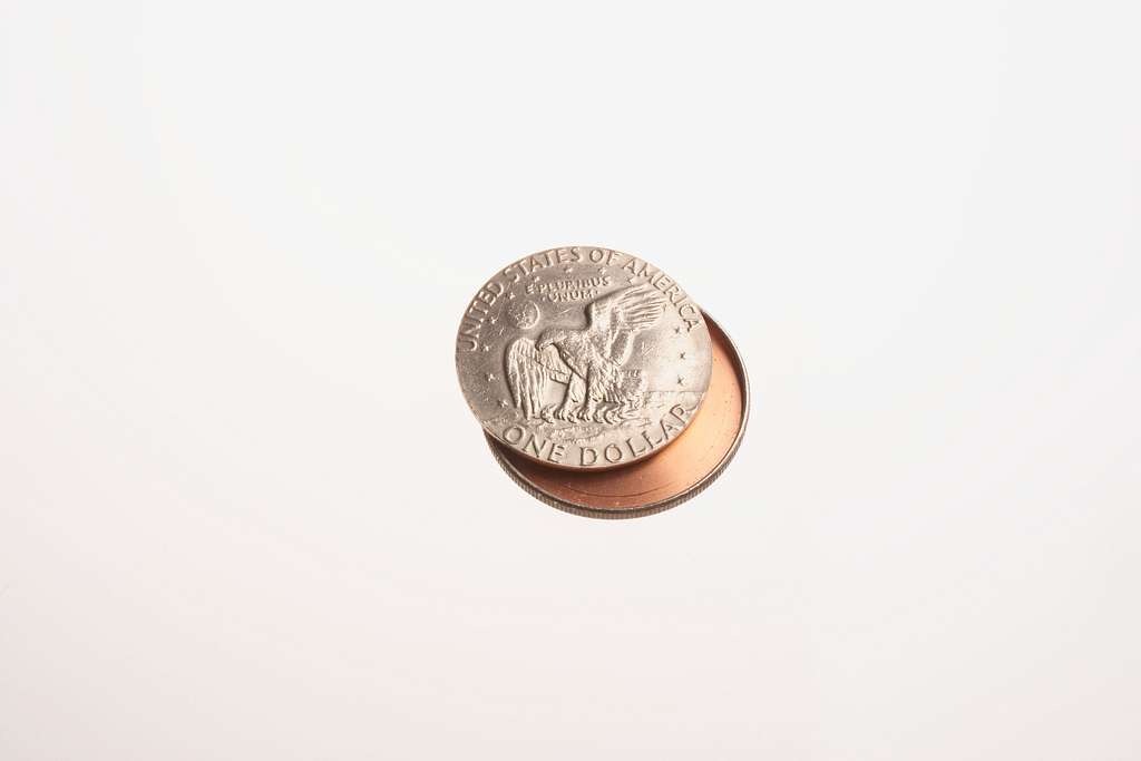 A silver American dollar coin, partially eclipsed by its shadow, is positioned against a plain white background. The coin features the image of an eagle and the words "United States of America" and "One Dollar.